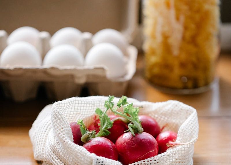 Organic fresh radishes in reusable bag placed on kitchen table near box with eggs
